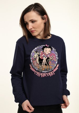 Marineblå sweatshirt med et grafisk motiv af en tegneserie-kvinde og en ged med et cirkulært design. Teksten nedenfor lyder "Stenbukken" i pink.