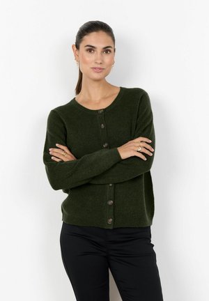 Frau mit dunkel zurückgebundenem Haar trägt eine dunkelgrüne, geknöpfte Strickjacke und schwarze Hose, steht mit verschränkten Armen vor weißem Hintergrund.