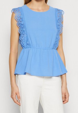 Woman wearing a sleeveless light blue top with floral lace trim and peplum hem, paired with white pants, standing against a plain background.