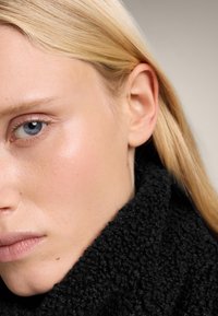 Prim-plan al unei fețe cu piele netedă, ochi albaștri și păr blond. Prezintă un guler texturat, de culoare neagră. Nu sunt vizibile accesorii sau modele.