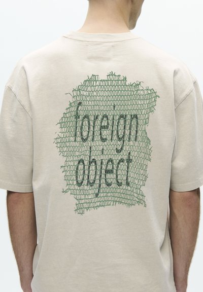 Personne portant un t-shirt beige avec un motif en filet vert et un texte noir "lovely object" au dos, tournée de dos à la caméra.
