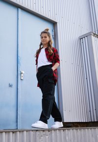 Chemise en flanelle bicolore rouge et noire, t-shirt blanc, pantalon cargo noir et baskets blanches. Debout près d'une porte en métal bleue.