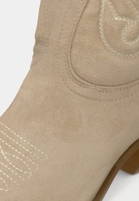 Μπεζ καουμπόικη μπότα από σουέτ με διακοσμητικό λευκό γαζί και διαμορφωμένη εγκοπή στον άξονα. Ξύλινο τακούνι και λεία υφή.