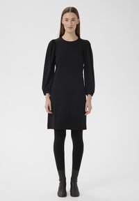 Rochie neagră cu mâneci lungi, umeri bufanți, croială dreaptă și textură moale, asortată cu colanți negri și cizme până la gleznă.