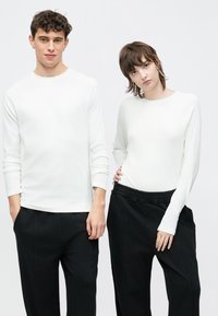 Kaks vaatemodelli kannavad valgeid, keha järgivaid pikki varrukatega toppe ümmarguse kaelusega, mida on kombineeritud lahtiste mustade pükstega, seisavad ühevärvilise tausta ees.