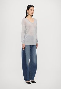 Un pull V en gris clair à manches longues, associé à un jean en denim à jambes larges avec des nuances de bleu variées, et des chaussures noires à bouts pointus.