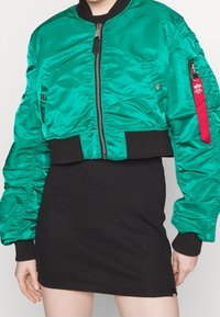 Groene bomberjack met een rits aan de voorkant, geribbelde manchetten en tailleband. Heeft een zak op de mouw met een rode label. Draagt over een zwarte jurk.