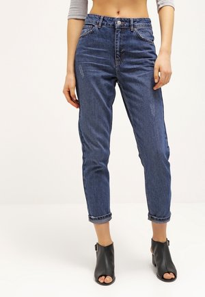 Kobieta w niebieskich, podwiniętych dżinsach z wysokim stanem i czarnych botkach z odkrytymi palcami, stojąca na jasnym, jednolitym tle.