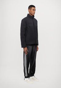 Puffer jacket in solid black with high collar, featuring a zippered chest pocket. Paired with glossy black track pants with white stripes.