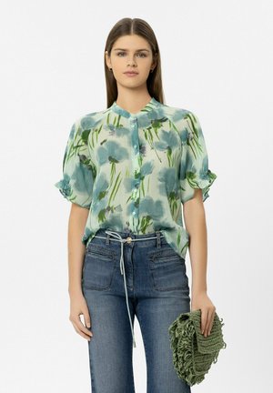 Frau trägt eine blau-grüne Blumbluse und Jeans, hält eine grüne Clutch mit Struktur vor einem schlichten weißen Hintergrund.