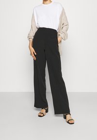 Black wide-leg trousers with a high waist, made of a smooth fabric. Paired with a white shirt and beige cardigan, complemented by black sandals.