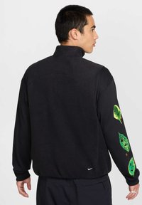 Black fleece jacket featuring a high collar and vibrant green leaf graphics on sleeves. Nike logo at the hem and a smooth texture.
