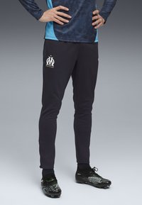 Pantaloni atletici de marină cu un design ajustat, având un logo alb pe partea stângă. Însoțiți de ghete de fotbal negre și verzi.