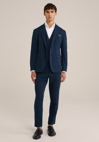 Abito tre pezzi blu navy su misura, caratterizzato da una giacca monopetto, un gilet e pantaloni slim, abbinato a una camicia bianca e mocassini neri.