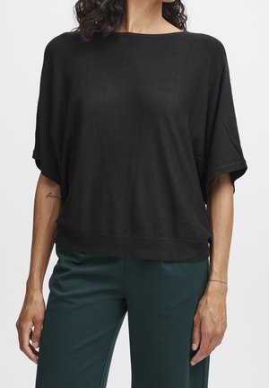 Haut noir à manches courtes avec un large col bateau et un tissu doux et léger. Associé à un pantalon vert, affichant une coupe décontractée.