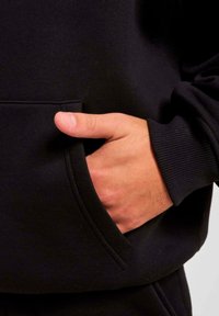 Felpa nera con tasca a marsupio e polsini a costine. Un primo piano di una mano appoggiata nella tasca mette in evidenza la texture e la vestibilità.