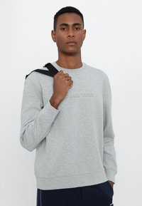 Sudadera gris de cuello redondo y mangas largas, con el logo en relieve de "Michael Kors" en el pecho. El modelo sostiene una correa de bolso.