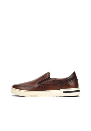 Zapato marrón de piel tipo mocasín con una suela de goma crema que contrasta. Presenta detalles de costura y paneles elásticos laterales para facilitar su uso.