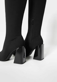 Botas negras hasta la rodilla hechas de un material suave y elástico. Presentan una cremallera lateral y un tacón geométrico, grueso y brillante.
