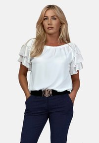 Biała bluzka z warstwowymi rękawami w falbanki, zestawiona z granatowymi spodniami o teksturowanym wykończeniu i czarnym paskiem z złotą okrągłą klamrą.