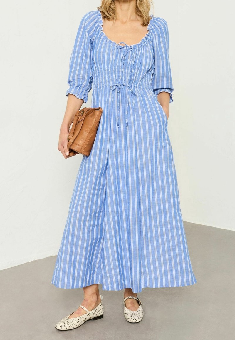 Femme portant une longue robe rayée bleu et blanc avec des manches trois-quarts, tenant un sac marron et portant des chaussures plates blanches tissées.