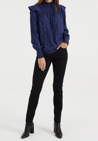 Žena v modré vzorované blůze s dlouhým rukávem, smockovaným límcem a manžetami, černých těsně přiléhavých kalhotách a černých kotníkových botách na podpatku, stojící.