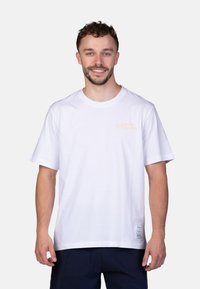 Witte katoenen T-shirt met korte mouwen, met een klein oranje logo op de borst en een label aan de onderzijde met verzorgingsinstructies.