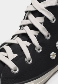 Μαύρο πάνινο αθλητικό παπούτσι με λευκά κορδόνια και μικρές λευκές διακοσμήσεις με λουλούδι μαργαρίτας κοντά στη σόλα.