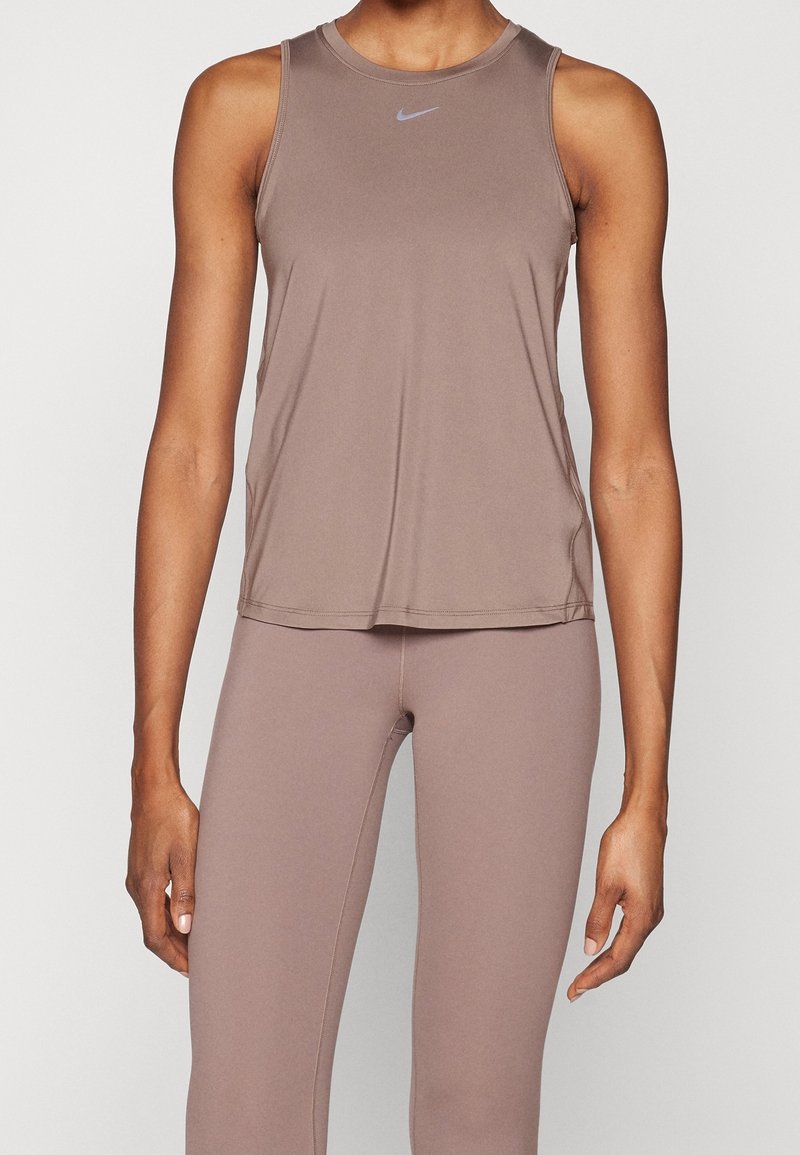 Personne portant un haut de sport sans manches mauve et un legging assorti, debout devant un fond clair uni.