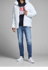 Giacca puffer bianca con zip frontale, indossata sopra un felpa scura con scritte rosse, abbinata a jeans blu e sneakers bianche.
