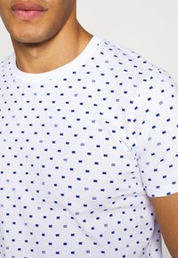 Hombre que lleva una camiseta blanca con pequeños patrones de bandera azul y "AB", vista desde el cuello hasta la mitad del torso.