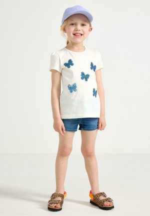 Ung pige iført en hvid t-shirt med sommerfugleprint, denimshorts, en lavendelfarvet kasket og sandaler med leopardprint, stående på en ensfarvet baggrund.