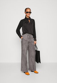 Schwarze Satinbluse mit Schlüssellochdetail, graue weit geschnittene Hose aus weichem Material, orangefarbene spitze Schuhe und eine schwarze Tragetasche.