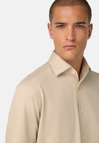 Camicia beige con bottoni e colletto strutturato, texture liscia e dettagli sulle cuciture laterali, posizionata su uno sfondo neutro.