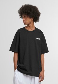 Black cotton oversized T-shirt with a crew neckline, featuring a white logo on the left chest and short sleeves. Smooth texture.