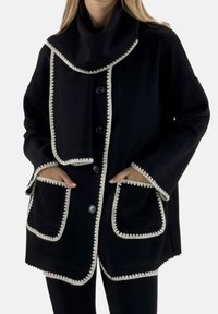 Zwarte jas gemaakt van een gestructureerde stof, met een hoge kraag, crèmekleurige rand langs de randen en grote voorzakken.