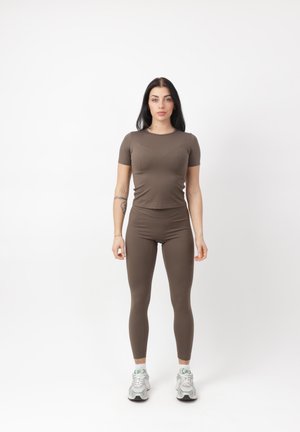 Mujer joven de pie mirando al frente, vistiendo una camiseta ajustada marrón y leggings, zapatillas blancas, sobre un fondo blanco liso.