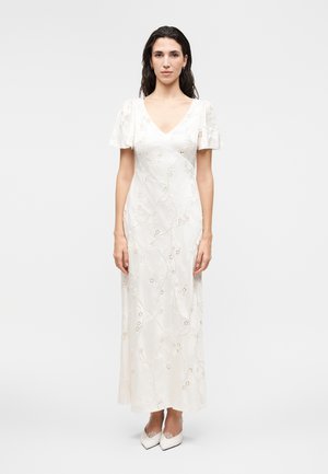 Γυναίκα με μακριά σκούρα μαλλιά που φοράει λευκό κεντητό φόρεμα με V-λαιμόκοψη και κοντά φουσκωτά μανίκια και άσπρα μυτερά παπούτσια, στέκεται σε μονόχρωμο φόντο.