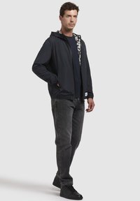 Leichte marineblaue Jacke mit Kapuze, ausgestattet mit einem Innenfutter im Tarnmuster, kombiniert mit dunkelgrauen Jeans und schwarzen Sneakers.