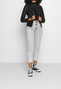 Black leather jacket with a zip front, worn over a fitted black top, paired with high-waisted light grey jeans and black sneakers.