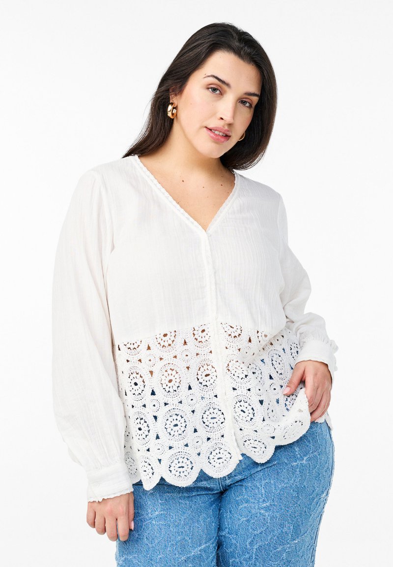 Vit blus med en texturerad överdel och en virkade spets underdel, med V-halsringning och långärmar, stylad med ljusblå denimjeans.