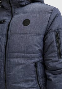 Padded jakna u plavobijelom materijalu s teksturiranim završetkom. Ima zatvaranje na patentni zatvarač, više džepova i okruglu oznaku na prsima.