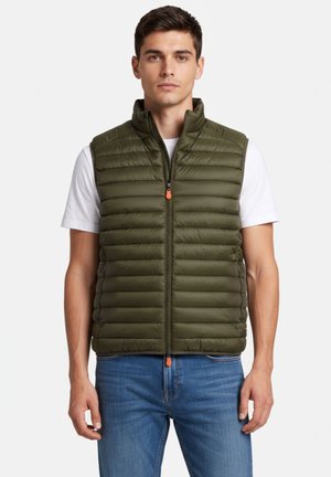 Giovane uomo che indossa un gilet trapuntato verde oliva sopra una maglietta bianca e jeans blu, in piedi davanti a uno sfondo semplice.