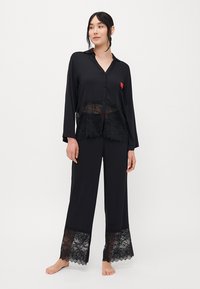 Woman standing barefoot wearing black lace-trimmed pajama set with buttons and collar, looking slightly to the side.