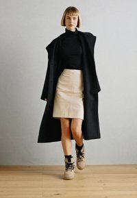 Palton negru peste un pulover pe gât negru, fustă din piele bej și adidași bej. Texturi netede, design elegant și palet�ă de culori neutre.