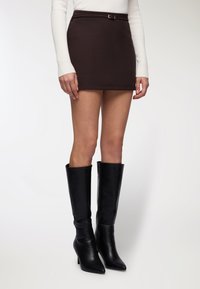 Donna che indossa una mini gonna marrone scuro, una maglietta bianca a maniche lunghe e stivali di pelle neri con punta a striscio, in piedi su uno sfondo bianco.