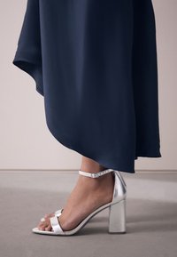 Pied portant une sandale argentée à talon bloc avec bride à la cheville, vue de côté, sous une longue robe fluide bleu foncé.