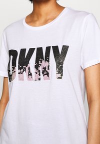 Person som bär en vit kortärmad t-shirt med stora svarta och rosa slitna "DKNY" bokstäver på framsidan.
