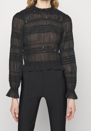 Femme portant un chemisier noir transparent à rayures horizontales et sequins, associé à un pantalon noir taille haute sur un fond uni.
