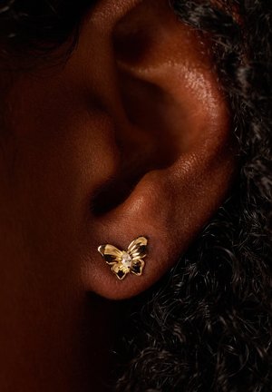 Boucles d'oreilles en forme de papillon en or avec une pierre précieuse transparente au centre, ailes détaillées, posées sur une peau foncée et des cheveux bouclés.